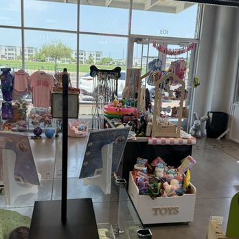 Pixie Sticks Dog Boutique - Boutique - The image shows the interior of a pet toy and accessory store with large glass front windows, allowing natural light to illuminate the space. Inside, there are shelves and displays filled with toys, accessories, and clothing items for pets, including plush toys, collars, and apparel. The store has a clean, open layout with a concrete floor and a view of parked cars and buildings outside through the windows. - Pet Groomers Near Me - Gilbert, Arizona