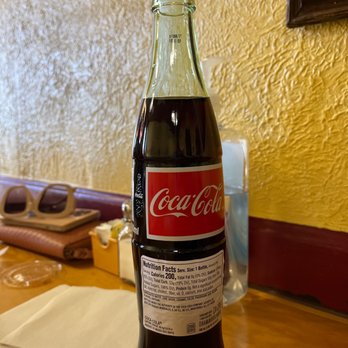 Papa Nachos - Real Coke!!!! - A glass Coca-Cola bottle filled with dark soda is positioned upright on a wooden restaurant table. The bottle has a red and white label with the Coca-Cola logo and nutrition facts. The background includes a textured yellow wall, a pair of sunglasses, napkins, and a tissue box, indicating a casual dining setting. - Mexican Near Me - Albuquerque, New Mexico