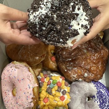Voodoo Doughnut - Dozen doughnuts - A box contains multiple donuts with different toppings, including sprinkles, icing, and crushed cookies, held by two hands. - Donuts Near Me - Denver, Colorado