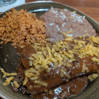 Cantina Laredo - Beef enchiladas  Beef - A ceramic plate holds a serving of Mexican food, including a portion of seasoned rice, refried beans, and a beef enchilada topped with shredded cheese and covered in brown sauce. The rice is orange-colored, and the beans are light brown. The enchilada is placed in the foreground, with the rice and beans positioned behind it. The plate is on a wooden table, with a glass of water and a napkin visible in the background. - Mexican Near Me - Bloomington, Minnesota
