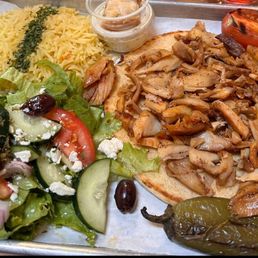 Chicken Doner plate - The image shows a Middle Eastern meal served on a metal tray, including yellow rice garnished with herbs, a fresh salad with cucumbers, tomatoes, olives, and feta cheese, grilled chicken pieces on flatbread, roasted tomato, and a roasted pepper. A small container of sauce or dip is also present.