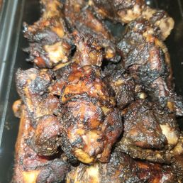 Jerk Wings - A collection of cooked chicken pieces with a dark, caramelized exterior, some areas showing charring, placed in a black plastic container. The chicken appears to be coated in a sauce or marinade that has caramelized during cooking.