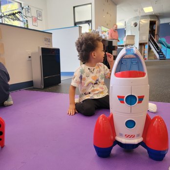 Over the Moon - A young child with curly hair is seated on a purple play mat inside an indoor playground, next to a plastic toy rocket with red, white, and blue colors. The child is wearing a shirt with various colorful patterns and appears to be engaged in play or observation. The background shows a man standing at a counter, with play structures, stairs, and wall decorations visible, indicating a space-themed indoor play area designed for children. - Indoor Playcenter Near Me - Cary, North Carolina