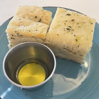Oceanview Diner - Two slices of garlic bread topped with herbs are placed on a blue ceramic plate. A small metal cup filled with olive oil is positioned next to the bread, likely for dipping. The presentation suggests a simple appetizer or side dish, typical of a dining setting that offers bread with olive oil. - Breakfast & Brunch Near Me - Norfolk, Virginia
