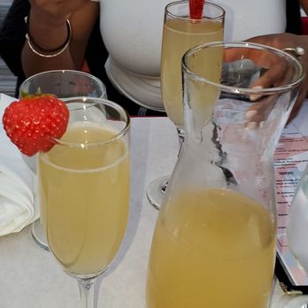 Patrick Steakhouse - Bottomless Mimosas - A woman with dark curly hair, wearing a white shirt and layered necklaces, is seated at a restaurant table. She is holding a smartphone in her right hand. On the table, there are three glasses of yellowish drinks, each garnished with a strawberry, and a glass pitcher filled with the same beverage. The setting appears to be a dining environment, possibly celebrating or enjoying drinks with friends. - Steakhouses Near Me - Brooklyn, New York