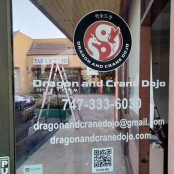 Burbank Printing - door decals - The image shows a glass storefront window with the Dragon and Crane Dojo logo at the top, depicting a dragon and a crane within a circular design. Below the logo, the business name, phone number, email address, and website are displayed in white text. A QR code is positioned at the bottom of the window, with a reflection of the street scene outside, including parked cars and neighboring storefronts. The window is part of a building with a wooden frame and a partial view of the interior on the right side. - Printing Services Near Me - Burbank, California