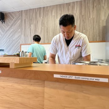 Chicago Sushi - A man wearing a white chef's uniform with 'CHICAGO RAMEN' embroidered on it is focused on preparing food behind a wooden counter in a restaurant. A customer in a light blue shirt stands with their back to the camera, facing the kitchen area. The setting features a wood-paneled wall and a sign on the counter that reads 'HEADS UP!! Sushi coming your way.' The environment appears to be a casual dining space with a minimalist design. - Japanese Near Me - Des Plaines, Illinois