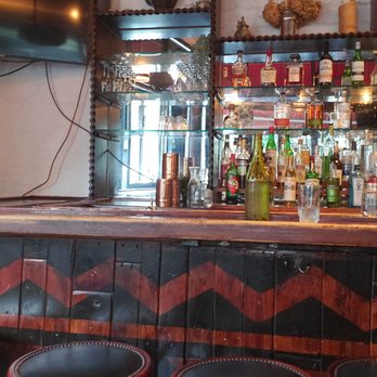 Cafe Rue Dix - Bar area - A bar area featuring a wooden counter with three red stools with black cushions. Behind the bar, there are shelves stocked with various liquor bottles, glasses, and decorative objects. The background includes a window with natural light and some wall-mounted decorations. The setting appears to be a restaurant or pub with a cozy, rustic interior. - French Near Me - Brooklyn, New York