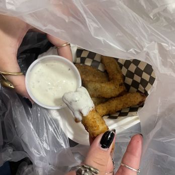 Fat Shack - fried pickles - A person with black nail polish and multiple rings holds a fried pickle dipped in a white creamy sauce. The pickle is partially covered in the sauce, and the basket below contains fried onion rings and chicken tenders. The basket is lined with checkered paper and is wrapped in a plastic bag, suggesting takeout or casual dining. - Sandwiches Near Me - Eugene, Oregon