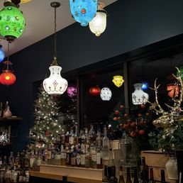 A bar interior features hanging lanterns of various colors and designs, a small decorated Christmas tree, and bottles of alcohol on the counter.