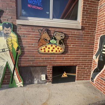 Old School Pizza Malta - Cardboard cutouts of Freddie Mercury, Elvis Presley, and cartoon characters are placed outside a pizza restaurant with brick walls. - Pizza Near Me - Malta, Illinois