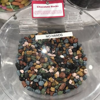 IT'SUGAR - Rock candy - A clear, round plastic container displays small, irregularly shaped chocolate rocks in various colors including brown, green, pink, black, and blue. A label on top reads 'Chocolate Rocks,' and a sign inside the container states 'NO HANDS.' The container is part of a candy display, with other candies visible in the background, including pastel-colored candies and black licorice. - Candy Stores Near Me - Phoenix, Arizona