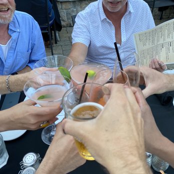 The Front Yard - Cheers - Several individuals are gathered around a table outdoors, raising their glasses in a toast. The drinks include cocktails with lime garnishes and a beer. The setting features patio furniture, a stone wall, and a building with windows and blinds in the background. The scene captures a social gathering during daytime or early evening. - Mediterranean Near Me - Ogunquit, Maine