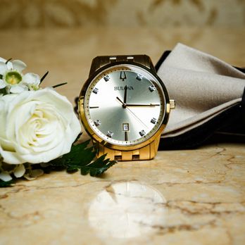 Capture Epic Moments - Fotografía de detalle que resalta un reloj dorado de estilo clásico, acompañado por una flor blanca y un moño formal, un conjunto típico en - Event Photography Near Me Capture Epic Moments - Fotografía de detalle que resalta un reloj dorado de estilo clásico, acompañado por una flor blanca y un moño formal, un conjunto típico en - Event Photography Near Me