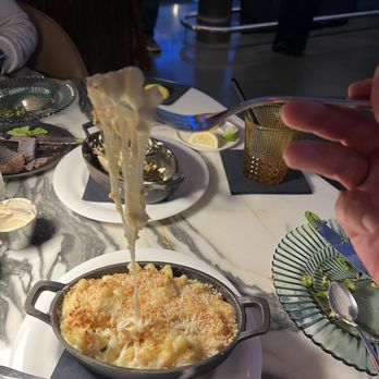 Sly Rooftop Bar - A fork with melted cheese is held above a small cast iron dish of baked pasta, with other dishes and drinks on a marble table. - Breakfast & Brunch Near Me - Washington, DC, District of Columbia
