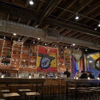 Toro - A bar interior with wooden shelving, bottles, and string lights, featuring a large colorful flag and a mural, with patrons seated at the bar. - Tapas Bars Near Me - Boston, Massachusetts
