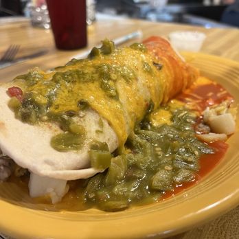 Duran Central Pharmacy - Breakfast Burrito - A breakfast burrito wrapped in a flour tortilla is placed on a yellow plate. It is topped with green chili sauce and melted cheese, with some red sauce visible on the side. The background shows a wooden table, a red drink, and a blurred restaurant setting. - Mexican Near Me - Albuquerque, New Mexico