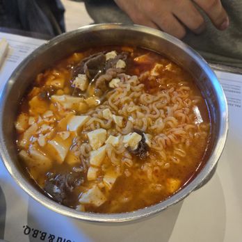 Sgd Dubu - A metal bowl contains spicy ramen with tofu, mushrooms, and curly noodles, placed on a restaurant table with chopsticks and a menu. - Korean Near Me - Madison, Wisconsin