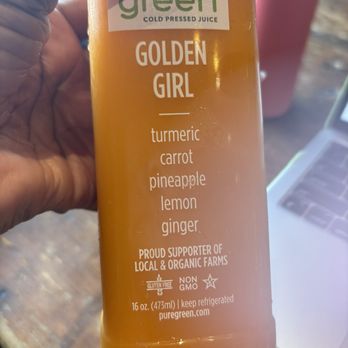Ground Central - Golden girl juice - A 16 oz bottle of Pure Green Cold Pressed Juice named 'Golden Girl' is shown, with ingredients including turmeric, carrot, pineapple, lemon, and ginger. The label indicates it is gluten-free, non-GMO, and supports local and organic farms. The bottle is held in a person's hand, with a blurred background including a pink cup and a laptop. - Coffee & Tea Near Me - Lynbrook, New York