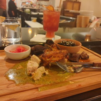 Mila's Mesa - Peruvian chicken. To die for! - A wooden serving board displays a portion of Peruvian chicken with green sauce, alongside black beans topped with cheese, and a small bowl of tomato salad. Two drinks, one clear with lemon and another red, are placed behind the board. The background shows a busy restaurant interior with staff and kitchen activity, indicating a dining setting focused on Latin cuisine. - Caterers Near Me - Albuquerque, New Mexico