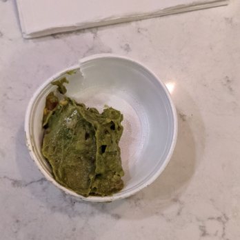 Estefani's Restaurant - Guacamole, barely. - A small, white disposable cup contains a portion of guacamole with a green color and chunky texture, placed on a light-colored marble surface. In the background, there is a white napkin or paper, partially visible. The image appears to be taken in a casual dining or takeout setting. - Breakfast & Brunch Near Me - Lynn, Massachusetts