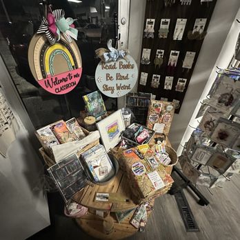 Whimsy & Wine - Unique Items - A round wooden display table in a gift shop features various jewelry items, books, and decorative signs, including a sign that reads 'Read Books Be Kind Stay Weird.' Behind the table, a window shows reflections of the shop interior and parking lot, with wall-mounted jewelry displays on the right. The shop interior has dark flooring and a mix of decorative and retail items. - Home Decor Near Me - Fraser, Michigan