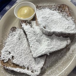 French Toast - Three slices of French toast are arranged on a white oval plate, generously dusted with powdered sugar. A small cup of butter or cream is placed on the plate. The dish is set on a checkered tablecloth, typical of a traditional diner or breakfast restaurant. The presentation emphasizes a classic breakfast item with a sweet topping.