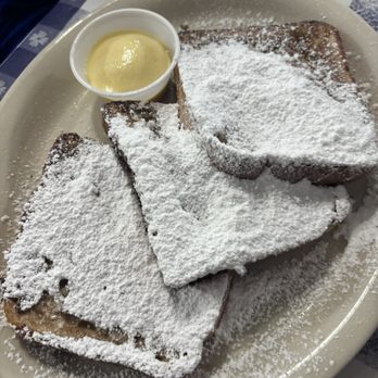 La Pines Cafe - French toast - Three slices of French toast are arranged on a white oval plate, generously dusted with powdered sugar. A small cup of butter or cream is placed on the plate. The dish is set on a checkered tablecloth, typical of a traditional diner or breakfast restaurant. The presentation emphasizes a classic breakfast item with a sweet topping. - Breakfast & Brunch Near Me - Slidell, Louisiana