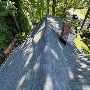Pro Touch Roofing  - The photograph shows an aerial view of a house's roof covered with gray asphalt shingles. A brick chimney with a metal cap and a vent are visible on the roof. The surrounding area includes tall trees with green foliage, a backyard with a deck, and a street with parked cars. The scene is illuminated by sunlight, casting shadows from the trees onto the roof. - Roofing Near Me