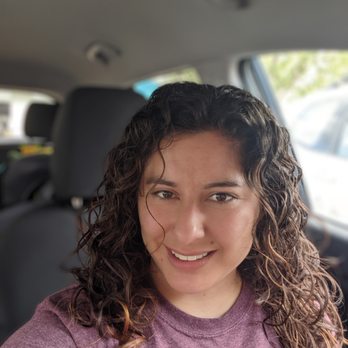 Roots Salon - A woman with curly hair and a purple shirt is taking a selfie inside a car, with the vehicle's interior and a blurred background visible. - Hair Salons Near Me - Albuquerque, New Mexico