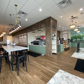 Corner Bakery Cafe - Lobby and Dining - The image shows the interior of a quick-service bakery with a seating area on the left, including tables and chairs. In the center, there is a coffee station with coffee dispensers and a display case. The right side features a mural of mountains and trees, along with a tiled floor section and a fire extinguisher. The ceiling has recessed lighting and hanging fixtures, creating a bright, welcoming atmosphere. - Bakeries Near Me - Denver, Colorado