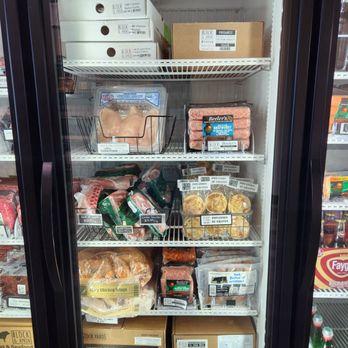 Block & Apron - Meats - A refrigerated display case contains various packaged meats, including chicken, steaks, and sausages, along with frozen side dishes like potatoes au gratin. The top shelves hold boxes of chicken and organic products, while the lower shelves feature ground beef patties and ribeye steaks in cardboard boxes. The case is part of a meat and seafood business, with clear labels and organized packaging. - Butcher Near Me - Gilbert, Arizona