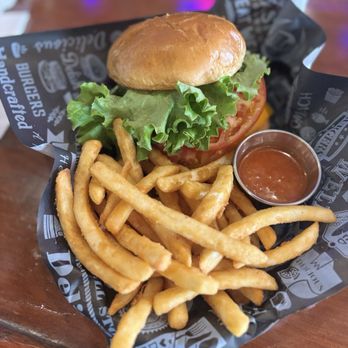 Sullivan's Cafe - Clucking Chicken (spicy chicken) - A burger featuring fresh lettuce, tomato, and a soft bun is presented on black and white branded paper, accompanied by a portion of French fries and a small cup of sauce, all placed on a wooden table. - Dive Bars Near Me - Naugatuck, Connecticut