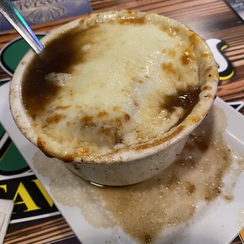Caffrey's Tavern Backyard - French Crock Of Onion Soup - A ceramic cup contains French onion soup topped with melted cheese, with some broth spilled onto the white plate beneath. The cup is on a wooden table with menus and utensils visible in the background, indicating a restaurant setting. - American Near Me - Forked River, New Jersey