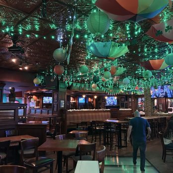 Jack & Gingers - The image shows a bar or pub interior with a ceiling decorated with umbrellas, paper lanterns, and string lights. The lighting is dim, with some illuminated screens visible in the background. The space includes tables, chairs, and a bar area, with a person walking across the floor. The decor suggests a celebration or themed event, with a lively and colorful ambiance. - Irish Pub Near Me - Austin, Texas
