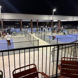 Outdoor courts - Night scene of multiple outdoor pickleball courts with players actively participating in games. The courts are enclosed by fences and illuminated by tall overhead lights. Spectators and additional players are visible in the background, some standing and some sitting. The area is covered by a large roof structure, and parked cars can be seen beyond the courts. The foreground includes a railing and chairs, indicating a viewing or seating area.