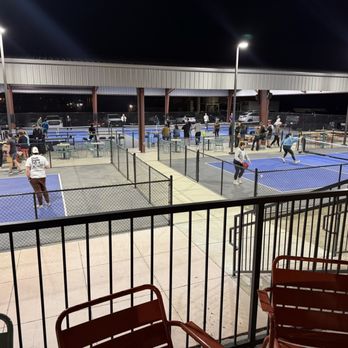 Moodswing - Outdoor courts - Night scene of multiple outdoor pickleball courts with players actively participating in games. The courts are enclosed by fences and illuminated by tall overhead lights. Spectators and additional players are visible in the background, some standing and some sitting. The area is covered by a large roof structure, and parked cars can be seen beyond the courts. The foreground includes a railing and chairs, indicating a viewing or seating area. - Venues & Event Spaces Near Me - Denver, Colorado