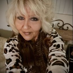 It's me coming out of Vons.  No long waits cashier  see that I needed help loading my groceries  .   I'm a happy camper. lol - A woman with blonde, curly hair and makeup is taking a selfie indoors. She is wearing a leopard print top and is smiling slightly. The background includes a bed with a decorative headboard and a wooden wall panel. The lighting is warm, and the focus is on her face and upper body.