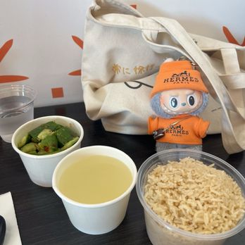 Cluck2Go - A table features a plastic container filled with cooked rice, a paper cup containing a light-colored soup, and a small bowl of chopped green vegetables. In the background, there is a beige tote bag with Japanese characters and a Hermes-themed plush toy dressed in orange with a Hermes hat and scarf. The setting appears to be casual, possibly in a cafe or restaurant, with a dark tabletop and a partially visible glass of water. - Singaporean Near Me - Pasadena, California