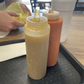 Los Cerritos - Salsas - Two squeeze bottles of condiments, one yellow and one red, are positioned on a black tray on a wooden restaurant table. The yellow bottle appears to contain mustard, and the red bottle likely contains ketchup. A large white drink cup with a straw is also on the table. In the background, there are blurred figures of people sitting at tables inside a restaurant, with natural light coming through the windows. - Mexican Near Me - Phoenix, Arizona