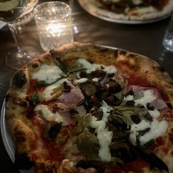 Lucia Rose - Capricciosa - A pizza featuring toppings such as black olives, artichokes, and ham is placed on a dark table. Surrounding the pizza are glasses of wine, a glass of water, and a candle. In the background, another pizza with similar toppings is partially visible. The setting appears to be a dimly lit restaurant or dining area. - Italian Near Me - Addison, Illinois