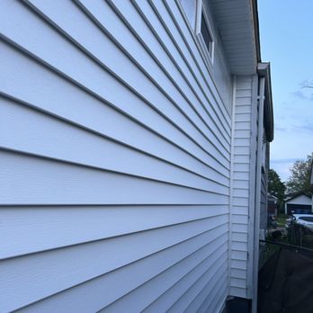 Morpho Carpentry - A house exterior features horizontal vinyl siding in a light color, with a small rectangular window near the top. The roof eaves extend outward, and part of the house's foundation is visible at the bottom. The sky is partly cloudy, and neighboring houses can be seen in the background. - General Contractors Near Me