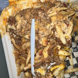 a tray of food - A serving of loaded fries with ground beef, melted cheese, and sauce is displayed on a tray. The fries are topped with cooked ground beef, shredded cheese, and some sauce, with a plastic fork inserted into the food. The tray has a checkered paper liner and some sauce residue around the edges.