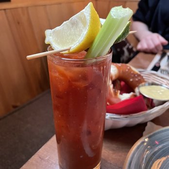 Stable DC - Bloody speck with smoked vodka - A tall, clear glass contains a Bloody Mary cocktail made with tomato juice and spices. The drink is garnished with a lemon wedge, a celery stalk, and a lime wedge, all skewered on a wooden pick. The setting appears to be a restaurant with wooden paneling in the background, and a basket of pretzels with mustard is visible nearby. - Modern European Near Me - Washington, DC, District of Columbia