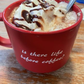 Fiddlestix Cafe - A red mug with the phrase 'is there life before coffee?' contains hot chocolate topped with whipped cream and chocolate syrup, placed on a wooden table. - Breakfast & Brunch Near Me - Cornwall, New York