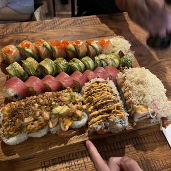 Domodachi - 98. EAT Therford Special Roll 102. Dragon Special Roll 105. My Love Tuna Special Roll 86. Spicy Salmon Crunchy Roll - A wooden platter holds various sushi rolls, including salmon, tuna, and specialty rolls, on a wooden table in a restaurant setting. - Sushi Bars Near Me - Rutherford, New Jersey