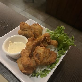 Shibui - Fried chicken - A white square plate holds several pieces of fried chicken, accompanied by a small container of creamy dipping sauce and a bed of fresh green lettuce leaves. The dish is presented on a dark wooden table in a restaurant setting, with a background showing tiled flooring and wooden furniture. - Japanese Near Me - Sunset, Florida