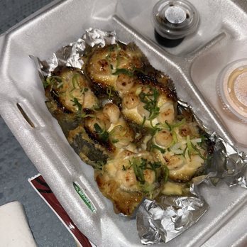 Yuzu Sushi - M-80 Roll - The image shows a foam takeout container with baked sushi rolls topped with scallions and a sauce, possibly eel or teriyaki. The sushi is placed on aluminum foil inside the container. Small containers of soy sauce and a dipping sauce are visible in the background. The setting appears to be a takeout or delivery meal, with the sushi arranged neatly for consumption. - Japanese Near Me - Chamblee, Georgia