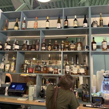 Beau's cafe - A person with long hair, wearing a green shirt, is seen working behind a bar counter. The background features a large shelving unit filled with various liquor bottles, including whiskey and wine. To the right, there is a coffee machine and cups, indicating a cafe or bar setting. The environment appears modern with a clean, organized layout. - Breakfast & Brunch Near Me - Lexington, Kentucky