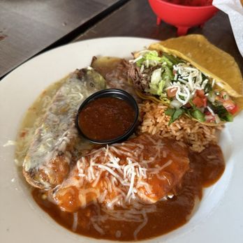 La Cueva Cafe - Combination Plate - A white plate holds a meal with Mexican and New Mexican cuisine, including rice topped with cheese, two enchiladas covered in sauce, and a taco filled with lettuce, tomato, and shredded cheese. A small container of salsa is placed on the plate. The setting includes a dark wooden table with a red bowl and a glass of water in the background, indicating a casual dining environment at La Cueva Cafe. - Mexican Near Me - Taos, New Mexico